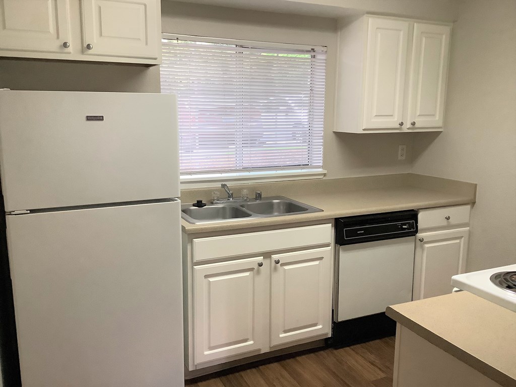 A white refrigerator stands in a kitchen with white cabinets and a window with blinds.