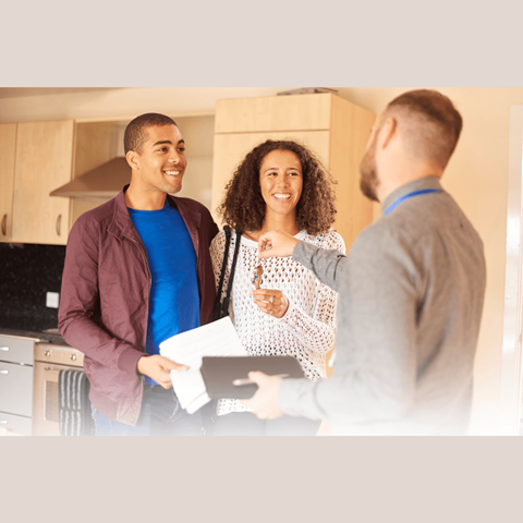 A man in a maroon jacket is holding a clipboard and appears to be taking notes while standing with two other people in a kitchen.