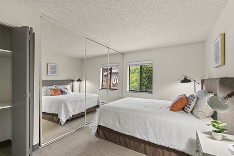 Apartments Near St. Joseph Medical Center, Tacoma, WA - The Orion - Bright Bedroom With Mirrored Closet Doors, Neutral Carpet, White Bedding, Bedside Lamps, and Window.