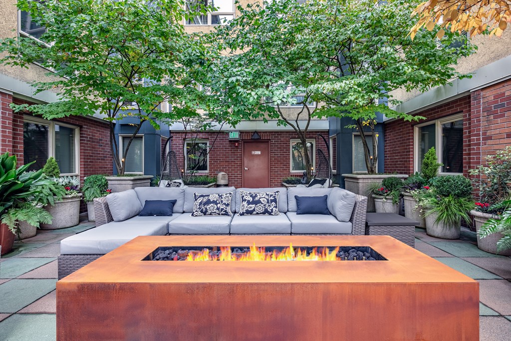 A fire pit in the middle of a patio surrounded by a white couch and potted plants.