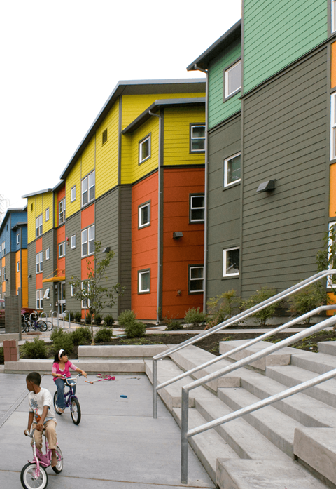 Two children riding bikes in front of a row of colorful buildings.