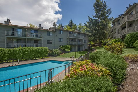 A swimming pool surrounded by a metal fence and bushes in front of apartment buildings.