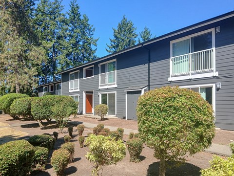 A grey building with a red door and windows surrounded by green bushes.