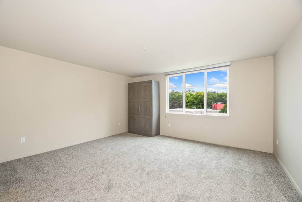 A room with a carpeted floor, a door, and a window showing a view of trees and a red building outside.