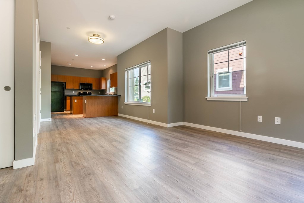 A spacious living room with wood flooring and a kitchen area in the background.