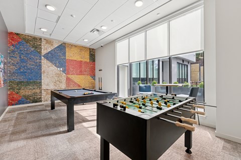 A foosball table is in the middle of a room with a pool table and a brick wall.