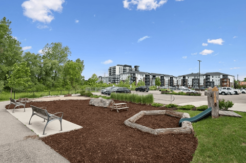 A park with a playground and benches in the foreground and apartment buildings in the background.