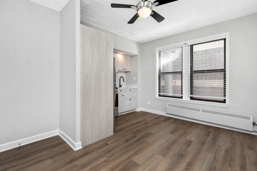 Living room with hardwood floors, white walls and a ceiling fan with a view into the kitchen