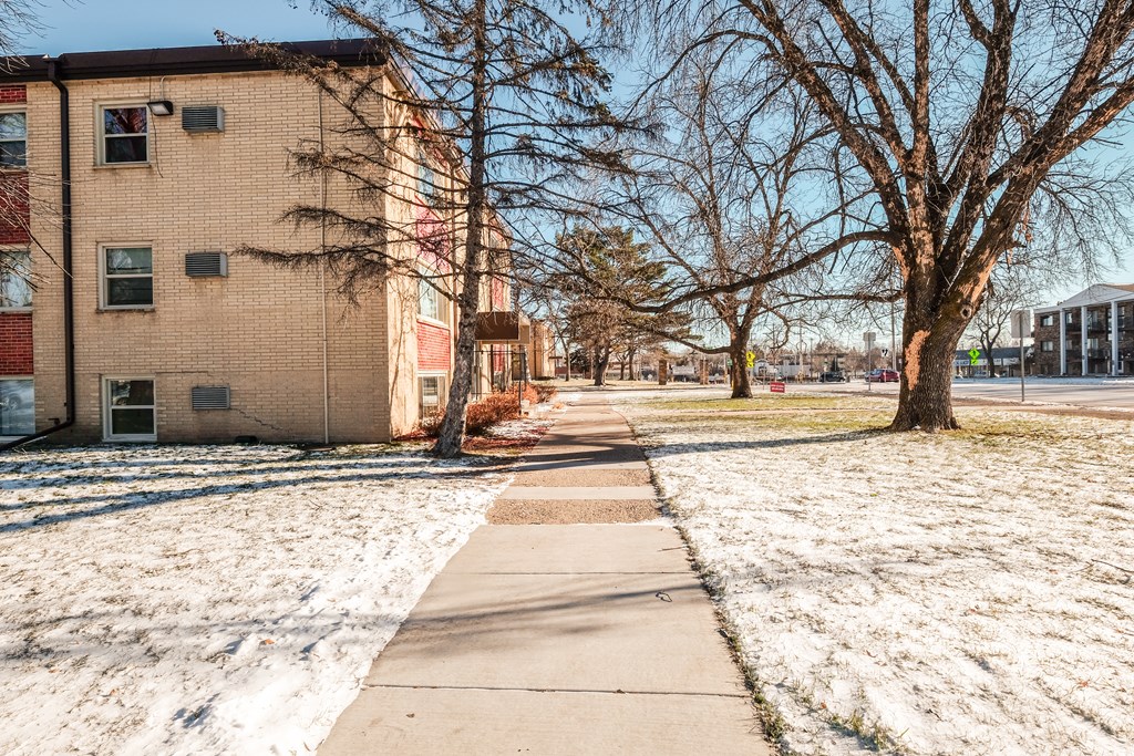 A snow-covered ground with a sidewalk and a tree in front of a brick building.