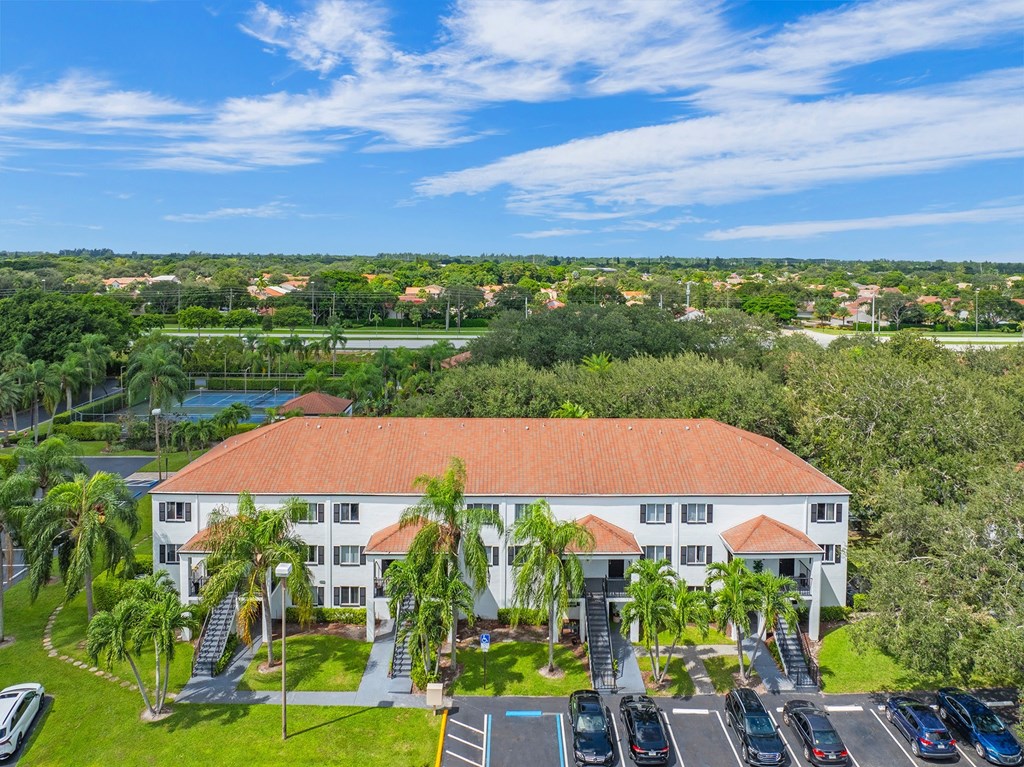 A large white building with a red roof is surrounded by greenery and has a parking lot in front.