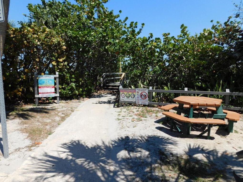A picnic table is located in a sandy area with a sign and a fence in the background.