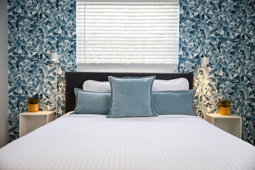 A bed with a white comforter and two blue pillows sits in a room with patterned wallpaper and two small tables with lamps.