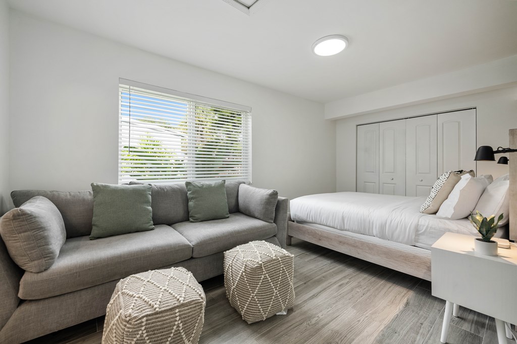A living room with a grey couch, a white bed, and a window with blinds.