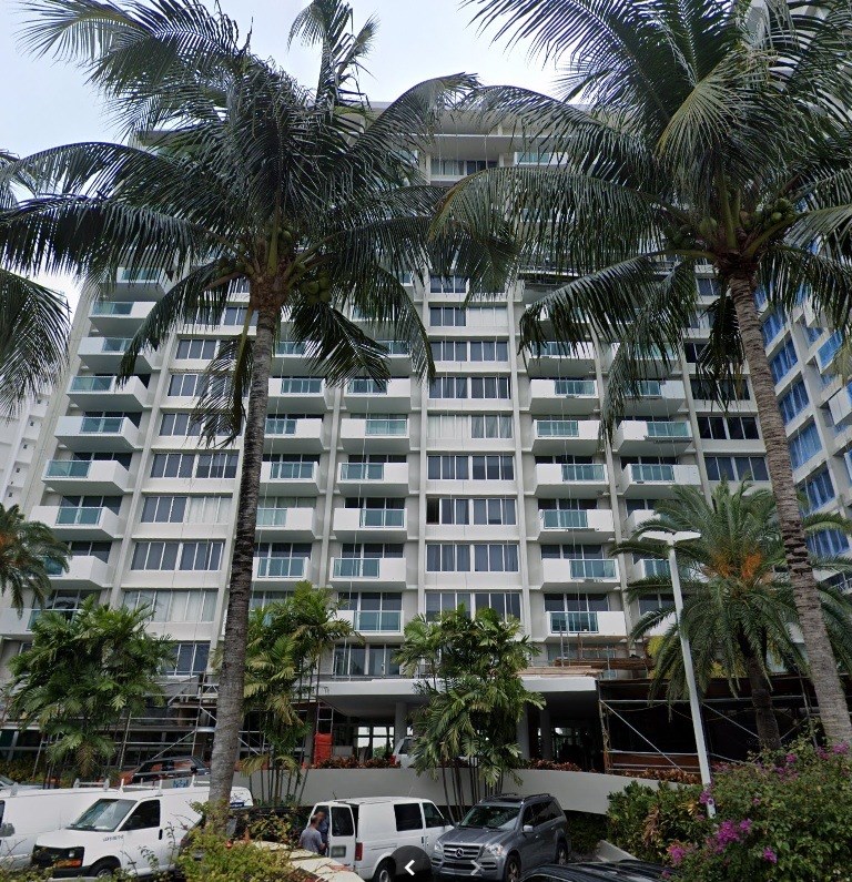 A large white building with balconies is surrounded by palm trees.
