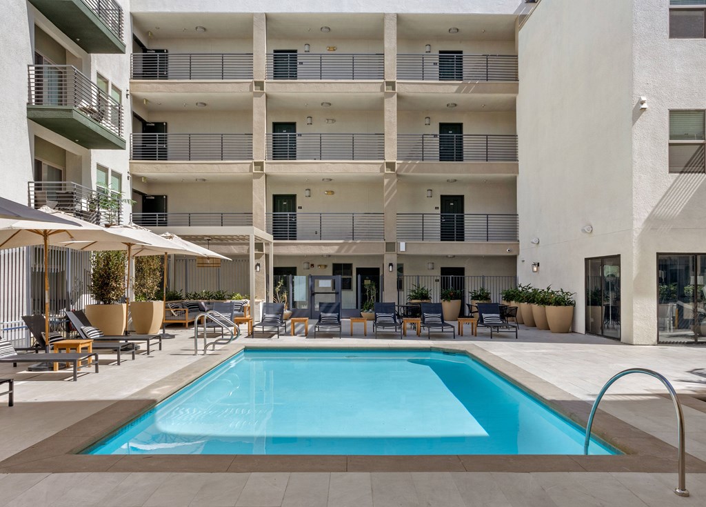 A large swimming pool is surrounded by lounge chairs and umbrellas in front of a multi-story apartment building.