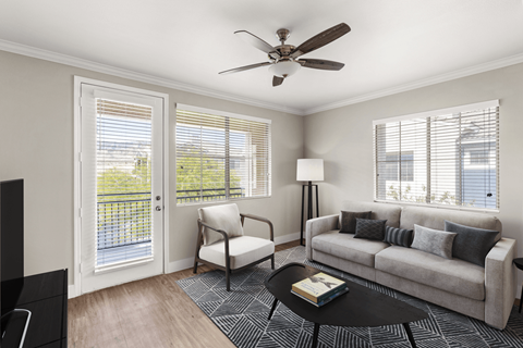 A living room with a grey couch, a black coffee table, a white chair, a ceiling fan, and a balcony with a railing.