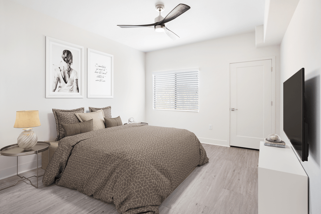 A bedroom with a bed, a ceiling fan, a lamp, a wall-mounted TV, and framed pictures on the wall.