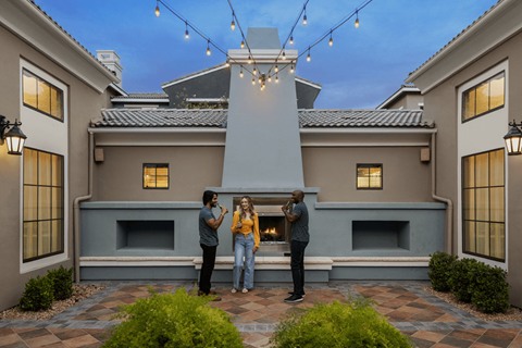 Three people stand in front of a grey house with a fireplace.