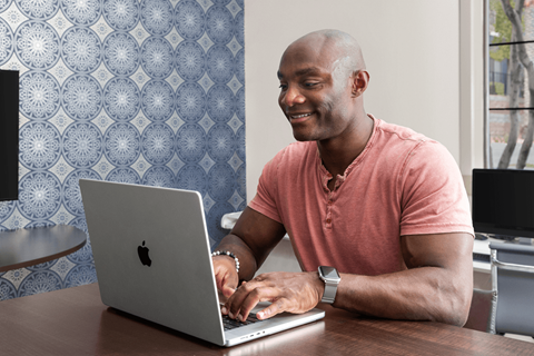 A man in a pink shirt is smiling while using a laptop.