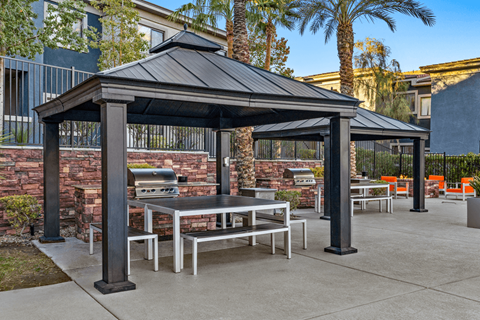 A black gazebo with a table and chairs is surrounded by a brick wall and palm trees.