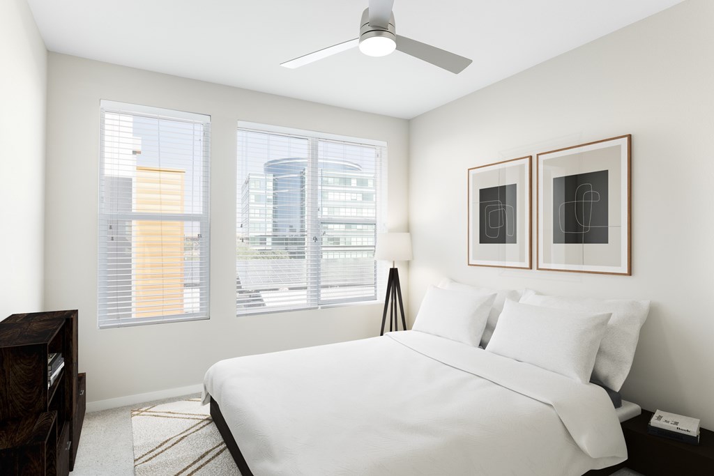 A bedroom with a large bed, a ceiling fan, and two windows with blinds.