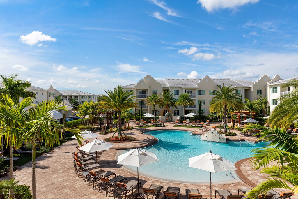 A large swimming pool surrounded by lounge chairs and umbrellas with a resort in the background.
