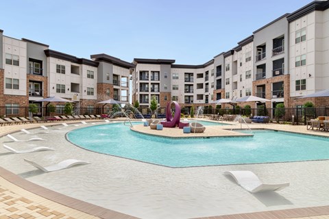 A large swimming pool surrounded by lounge chairs and umbrellas in front of apartment buildings.