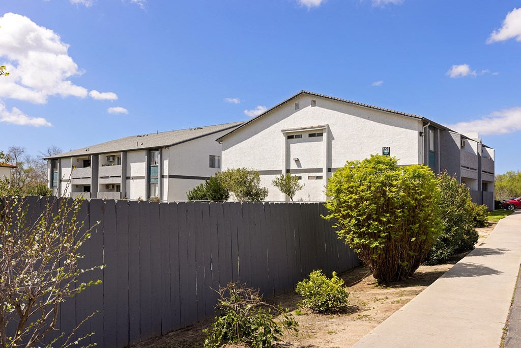 A grey building with a white door and windows is surrounded by a grey fence and green bushes.