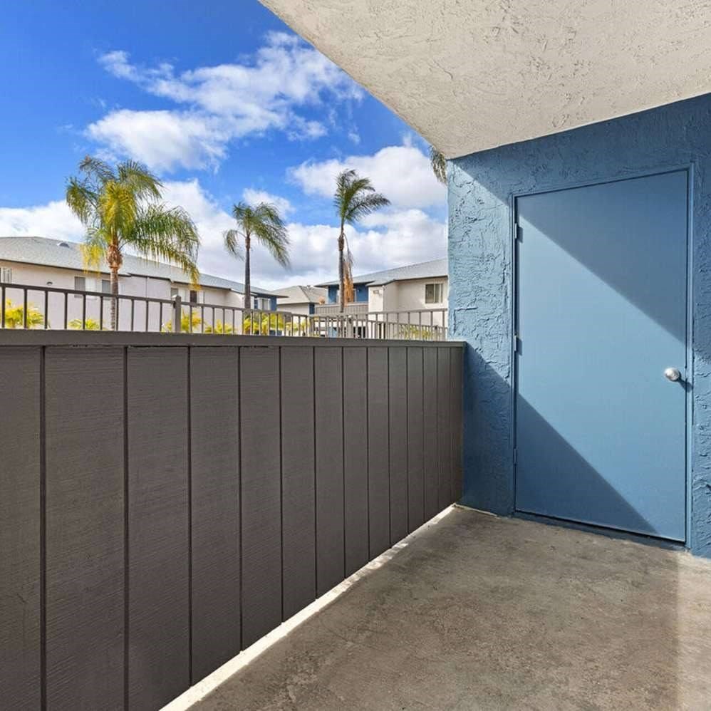 A blue door is on the right side of a wall with a balcony in the background.
