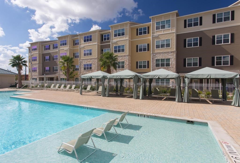 A swimming pool with sun loungers and umbrellas in front of a multi-story apartment building.