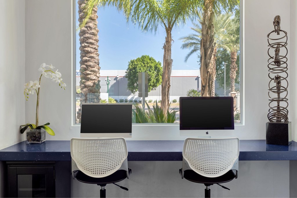 Two computer monitors are on a desk with a view of palm trees outside the window.