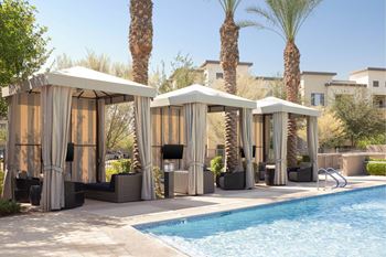 Four palm trees are behind a pool with four sun shelters.