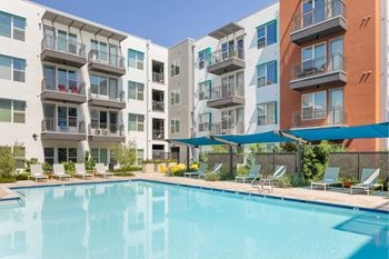 A swimming pool is surrounded by lounge chairs and umbrellas in front of apartment buildings.