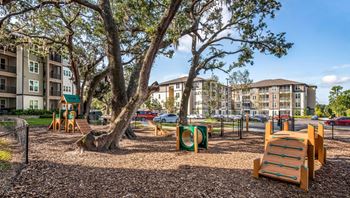 A playground with a slide, swings, and a tree in the foreground with apartment buildings in the background.