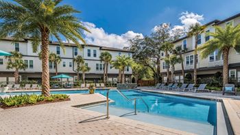 A swimming pool surrounded by palm trees and lounge chairs in front of apartment buildings.