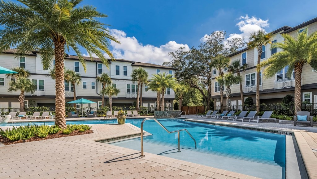 A swimming pool surrounded by palm trees and lounge chairs in front of apartment buildings.