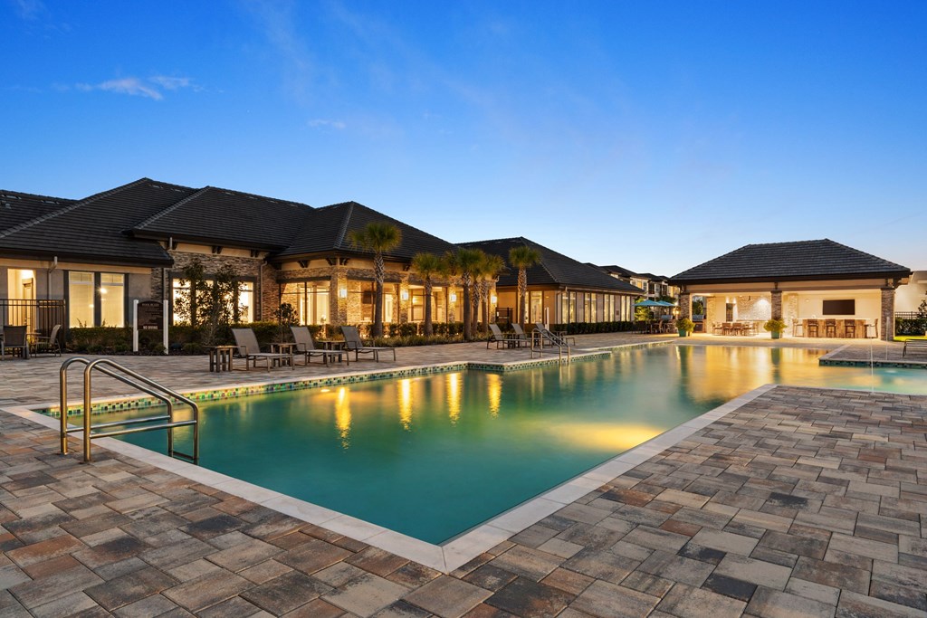 A large outdoor swimming pool surrounded by a stone patio and a building with a black roof.