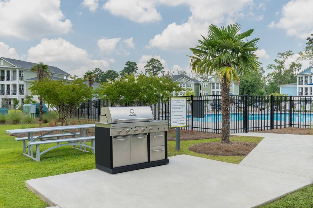 A BBQ grill is in the middle of a concrete area with a pool and palm tree in the background.