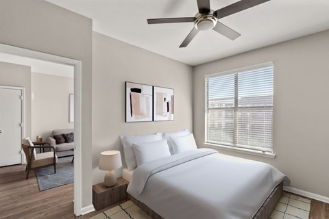 A bedroom with a bed, a chair, a table with a lamp, and a ceiling fan.