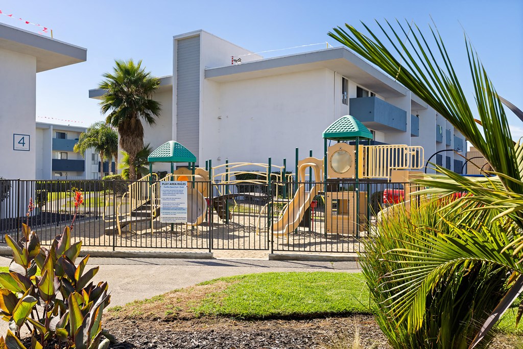 A playground with a slide and a green canopy is surrounded by a fence and a building with a number 4 on it.