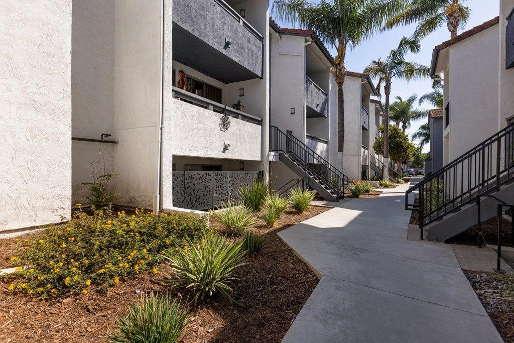 A concrete pathway leads to a building with a balcony and a small garden with yellow flowers.
