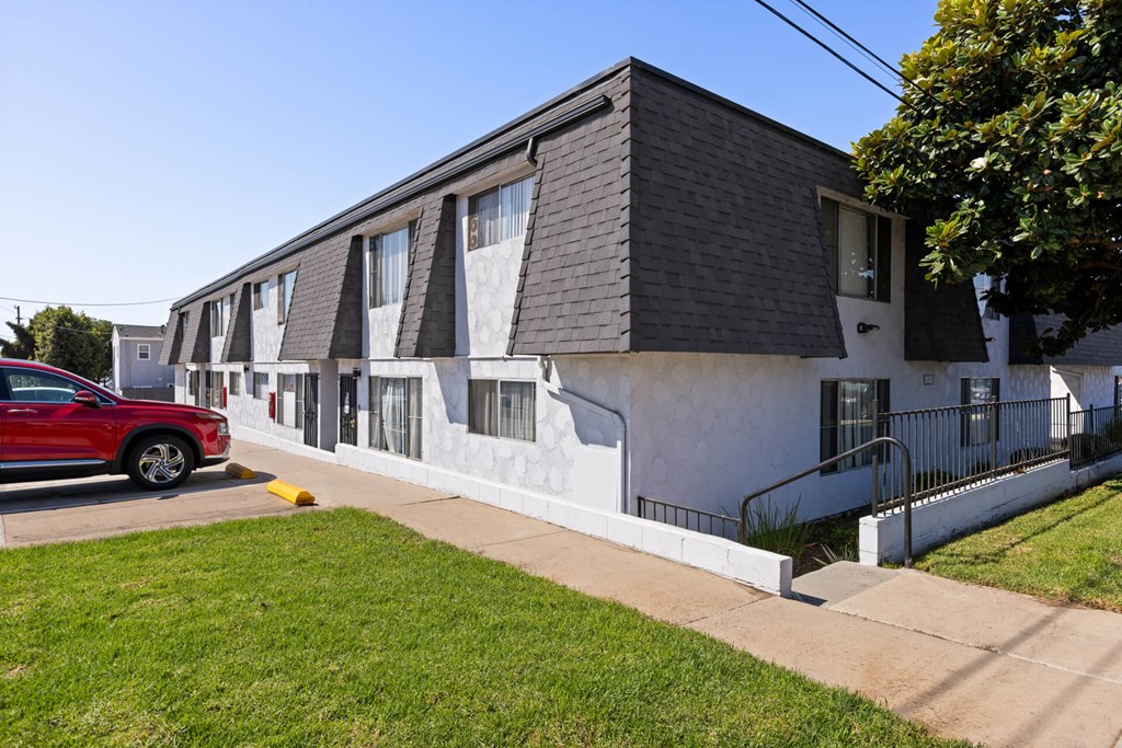 A red car is parked in front of a white building with a black roof.