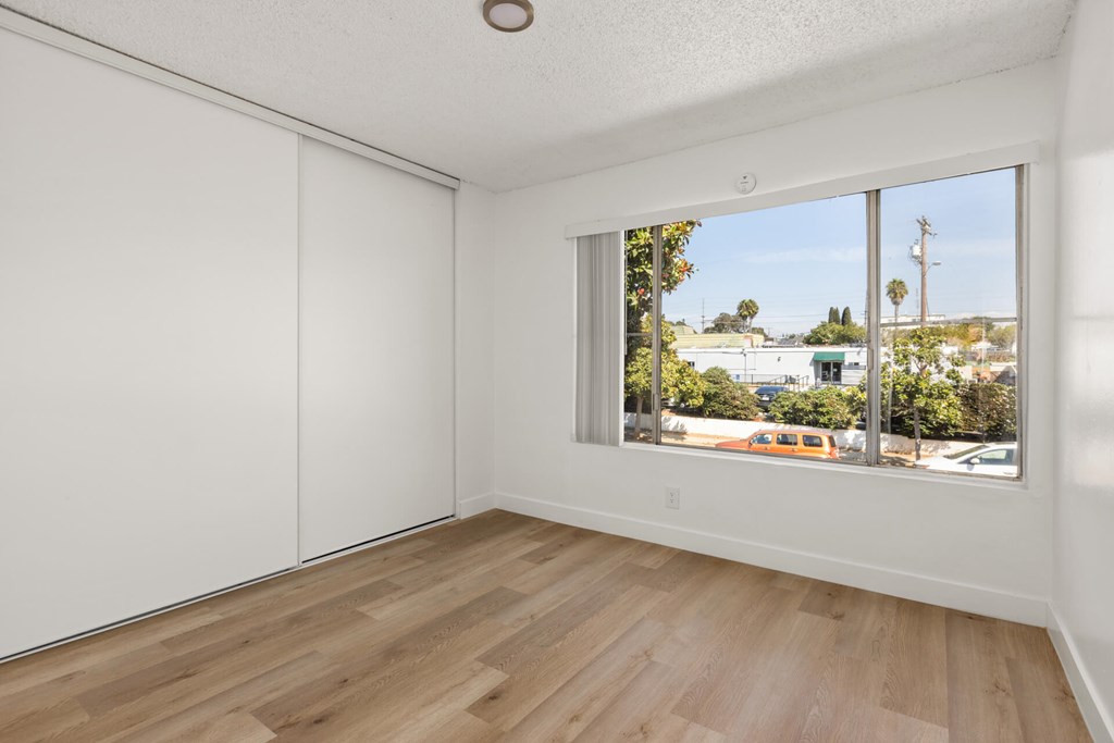A room with white walls and wooden flooring with a window showing a view of a street with cars and trees.