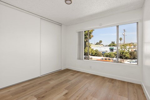 A room with white walls and wooden flooring with a window showing a view of a street with cars and trees.