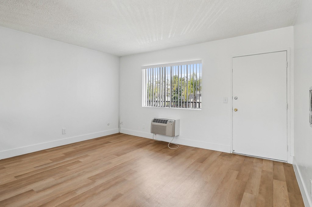 A room with wooden floors and white walls, a window with blinds, and a white door.