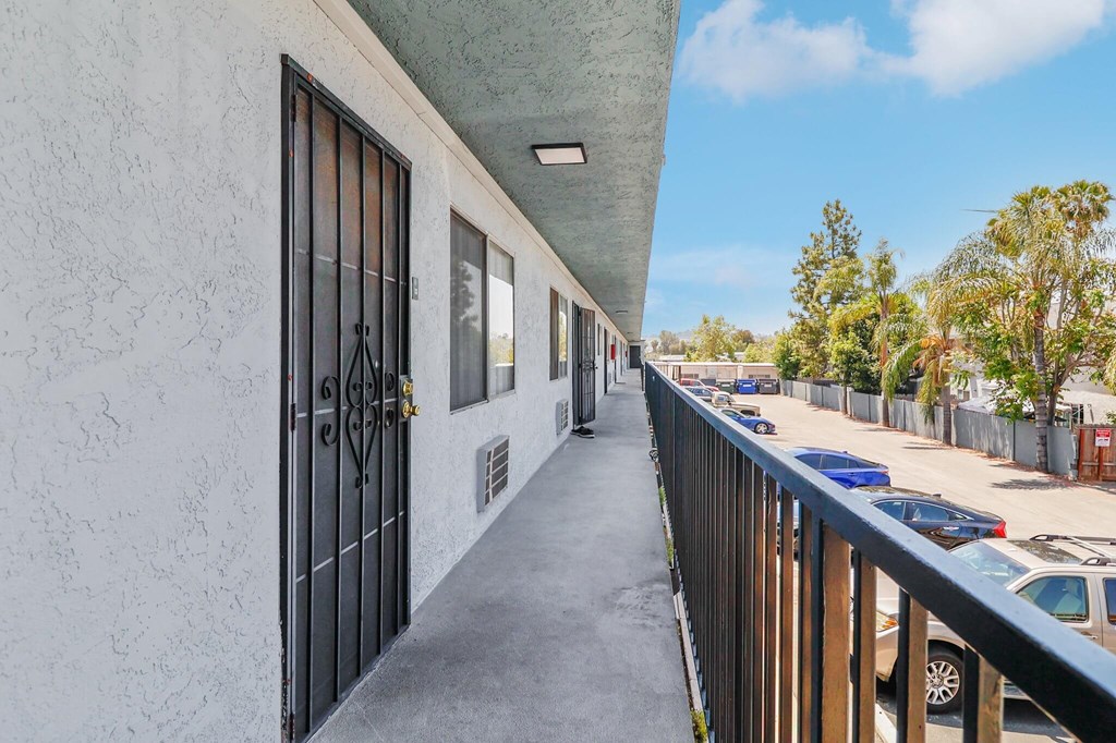 A long, narrow balcony with a metal railing and a white wall with a black gate.