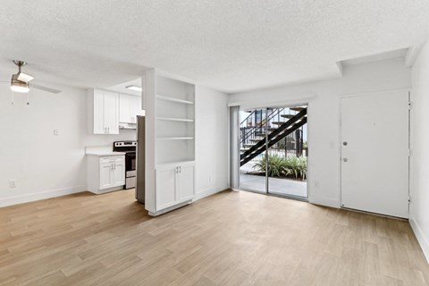 A room with a wooden floor and white walls, with a kitchen area and a staircase visible through the glass door.