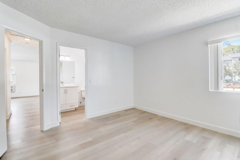 A room with white walls and wooden flooring, with a window showing a building outside.