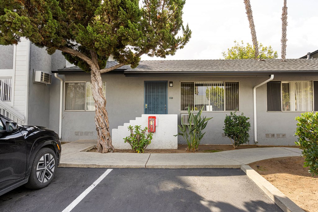 A black car is parked in front of a two-story house with a tree in front of it.