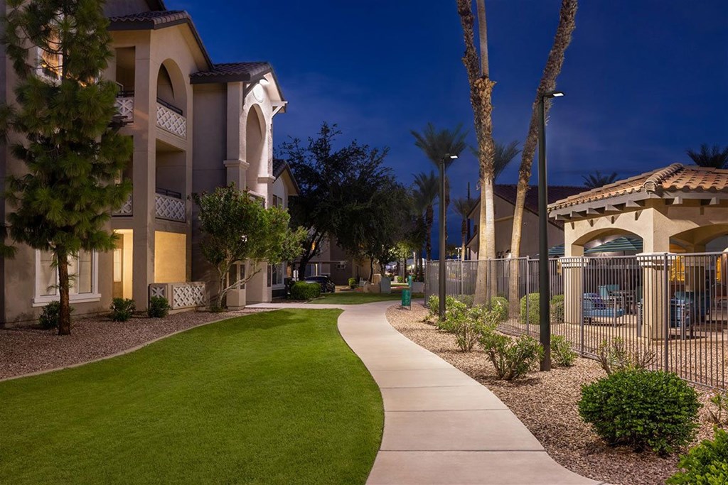 A well-lit pathway leads through a landscaped area in front of a building.